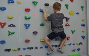 ćwiczenie pośladków na ściance wspinaczkowej. Dziecko stoi przodem na ściance w półprzysiadzie, kończyny dolne w rotacji zewnętrznej.