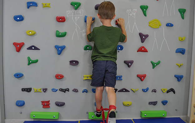 ćwiczenia pośladków na ściance wspinaczkowej. Dziecko stoi wyprostowane przodem na ściance wspinaczkowej. Wokół kostek zawiązana taśma elastyczna.