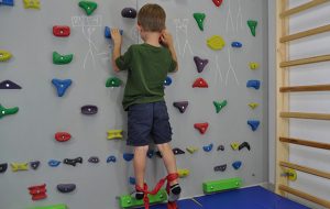 ćwiczenia pośladków na ściance wspinaczkowej. Dziecko stoi wyprostowane przodem na ściance wspinaczkowej. Wokół kostek zawiązana taśma elastyczna.