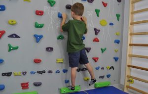 ćwiczenia pośladków na ściance wspinaczkowej. Dziecko stojąc przodem do ścianki wspinaczkowej unosi prostą nogę w bok.