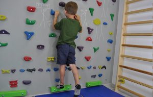 ćwiczenia pośladków na ściance wspinaczkowej. Dziecko stojąc przodem do ścianki prostuje nogę w tył napinając pośladek.