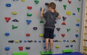 ćwiczenie na odstające łopatki i plecy okrągłe. Dziecko stoi przodem na ściance wspinaczkowej. Kończyny górne zgięte trzymają chwyty.