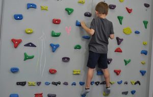 ćwiczenie pośladków na ściance wspinaczkowej. Dziecko stojąc przodem do ścianki z wyprostowanymi kolanami.