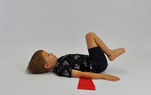 ćwiczenia na mięsnie brzucha. Dziecko leży na plecach, nogi ugięte, stopy na podłodze, pod odcinkiem lędźwiowym taśma elastyczna. Dziecko odrywa nogi i przyciąga je do brzucha rodzic pociąga za taśmę próbując ją wydostać