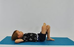 ćwiczenia na mięśnie brzucha. Dziecko leży na plecach, ręce na karku, stopa spoczywa na kolanie jak na zdjęciu