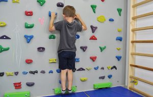 : ćwiczenia na stopy płasko-koslawe. Dziecko stojąc na ściance na palcach, schodzi piętami nisko w dół rozciągając łydki