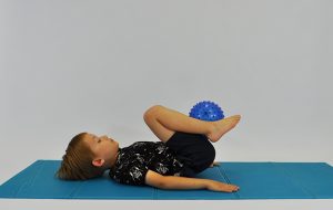dziecko w leżeniu tyłem trzymając między stopami piłkę, odrywa stopy i kolana przyciąga do klatki piersiowej