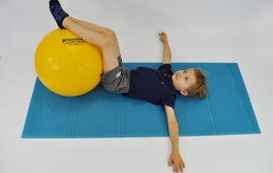 dziecko w leżeniu tyłem nogi trzyma na piłce, napina brzuch i skręca kolana z piłką w obie strony