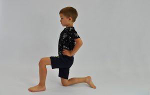 ćwiczenie rozciągające staw biodrowy. Dziecko klęczy na jednej nodze, ręce na biodrach, dziecko idzie miednicą w przód rozciągając biodro nogi opartej na kolanie