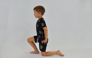ćwiczenie rozciągające staw biodrowy. Dziecko klęczy na jednej nodze, ręce na biodrach, dziecko idzie miednicą w przód rozciągając biodro nogi opartej na kolanie