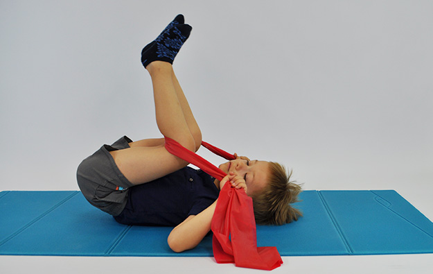 ćwiczenia rozciągające kręgosłup lędźwiowy. Dziecko leży na plecach w rękach trzyma taśmę elastyczną zaczepioną pod kolanami. Dziecko przyciąga kolana w raz z taśmą do klatki piersiowej