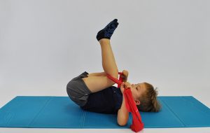 ćwiczenia rozciągające kręgosłup lędźwiowy. Dziecko leży na plecach w rekach trzyma taśmę elastyczna zaczepiona pod kolanami