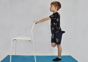 ćwiczenie rozciągające mięsień prosty uda. Dziecko stoi przodem do krzesła. Prawa ręka trzyma oparcie krzesła, lewą ręką przyciąga zgiętą piętę do pośladka rozciągając przednią stronę uda.