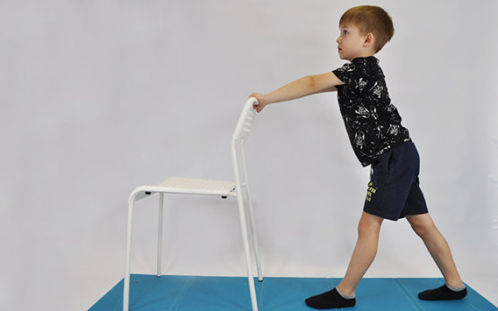 ćwiczenie rozciągające mięśnie łydek. Dziecko stoi przodem do krzesła (ściany stołu), lewa noga z przodu ugięta w kolanie, prawa z tyłu wyprostowana. Dziecko przenosi ciężar ciała na lewą nogę rozciągając prawą łydkę i nie odrywając jej od podłogi.